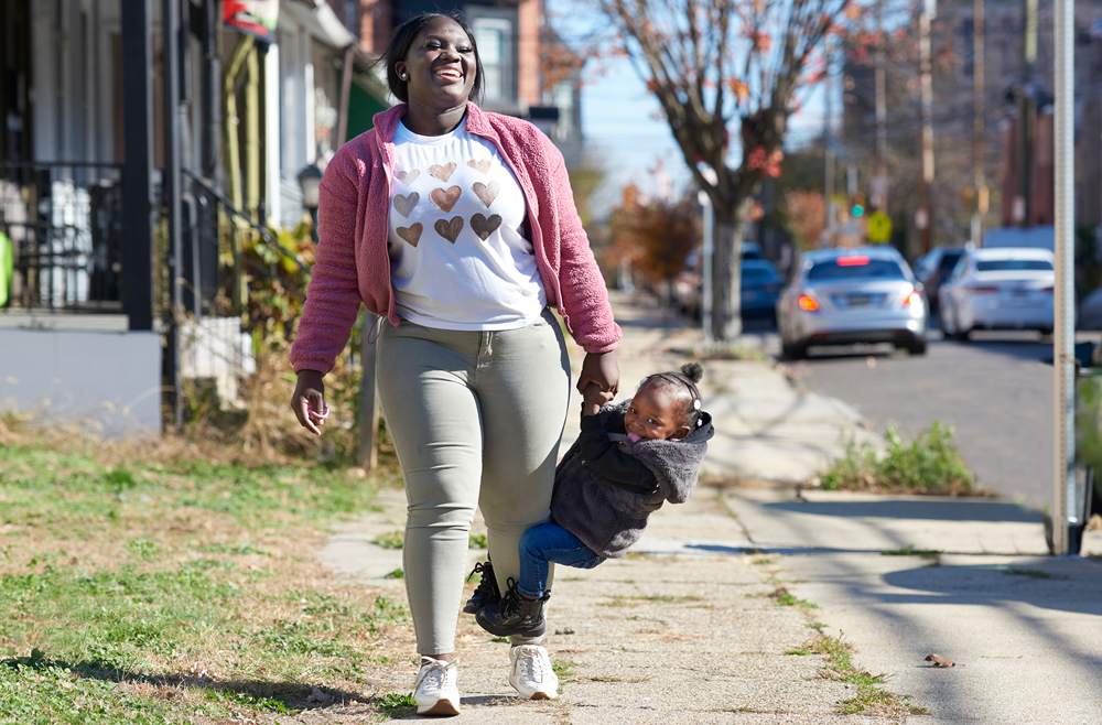 A mother, Talicia Williams, walks on a sidewalk while her 2-year-old daughter swings from her hanging left arm. 